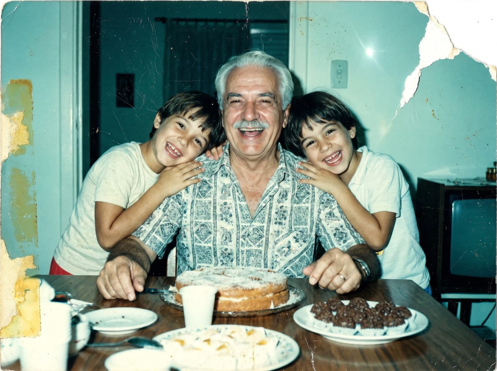 Foto de aniversário dos anos 1980, desbotada e manchada: um senhor de bigode abraçado por dois netos à mesa